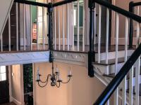 Two-story stair hall with classic wood balustrade, black handrails, and decorative trim, featuring a central iron chandelier and warm-toned walls, leading to bedrooms with painted walls and natural light.